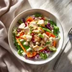 An overhead photo of a chicken salad featuring grilled chicken, cherry tomatoes, red cabbage, broccoli, carrots, and bell peppers, all drizzled with a rich dressing.